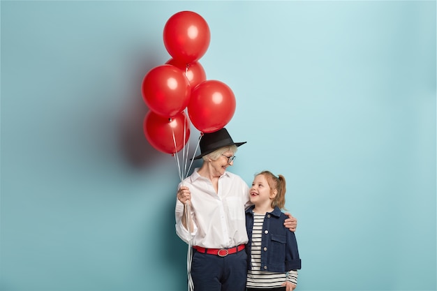 Happy caring grandmother holds bunch of red air balloons, congratulates granddaughter with birthday