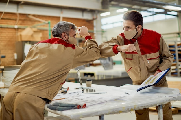 Happy carpenters elbow bumping while working in a workshop during coronavirus pandemic