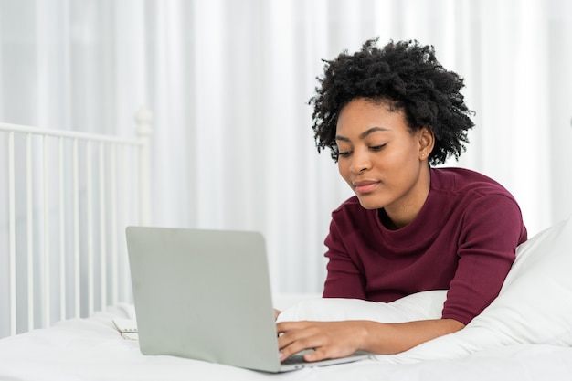 행복한 캐주얼한 아름다운 미국 아프리카 여성이 집에 있는 침대에 누워 노트북 컴퓨터를 하고 있습니다.