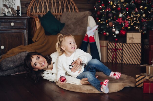 Happy casual young mother cute smiling daughter having fun at Christmas tree background full shot. Beautiful family feeling love and positive emotion enjoying Xmas decoration surrounded by snowflakes