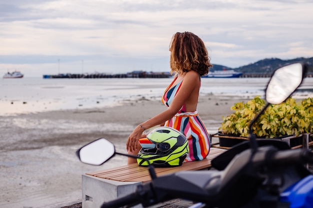 オートバイのヘルメットと休暇でカラフルな夏のドレスで幸せな白人バイカーの女性