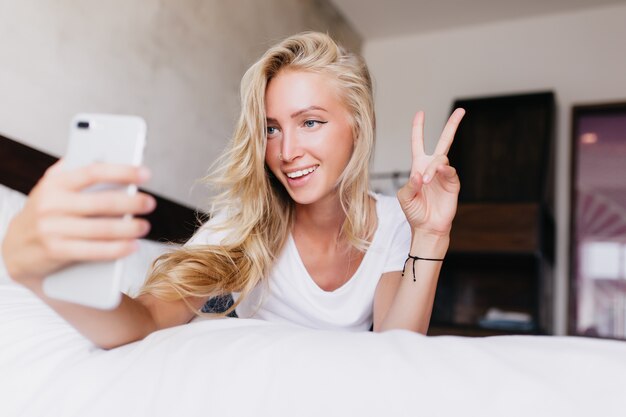 ピースサインで自分撮りを作る幸せな白人の金髪の女性。寝室で身も凍るように写真を撮る素敵な女性の室内撮影。