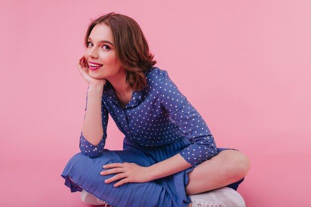 Счастливая кавказская девушка в синем наряде сидит на полу со сложенными ногами. Крытый выстрел элегантной леди брюнетки, улыбающейся на розовой стене.