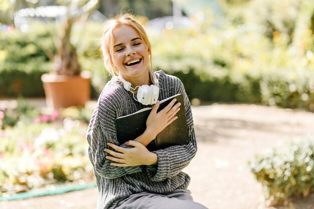 Free photo happy caucasian girl with headphones laughing in autumn garden blonde female student posing with book and smiling