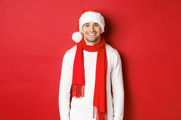 Счастливый кавказский парень празднует Рождество и улыбается в шарфе Санта-Клауса и белом свитере ...
