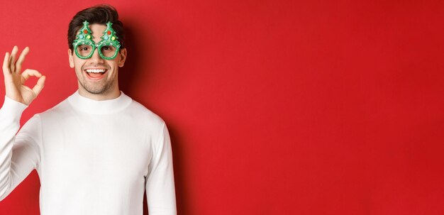 Happy caucasian guy wishing merry christmas, celebrating winter holidays, showing okay sign and smiling pleased, standing over red background.