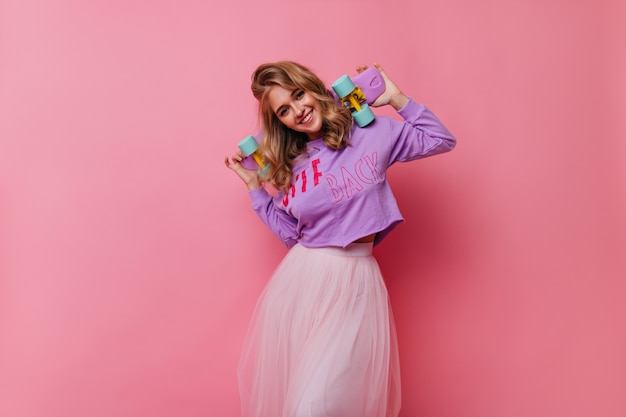 Happy caucasian lady having fun. Enthusiastic fair-haired girl with skateboard standing on pink.