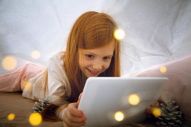 Счастливая кавказская маленькая девочка во время видеозвонка или обмена сообщениями с Дедом Морозом с помощью ноутбука и домашних устройств