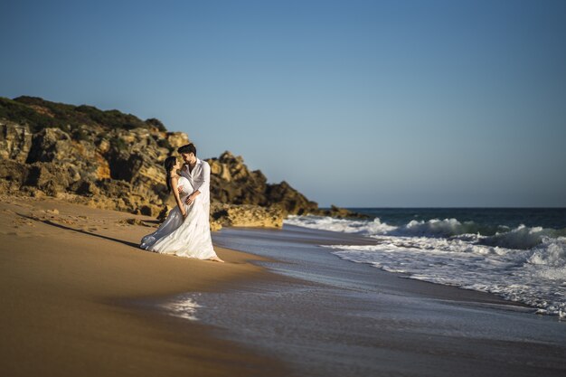 결혼식 사진 촬영하는 동안 해변에서 흰색 포옹을 입고 행복 백인 사랑하는 부부