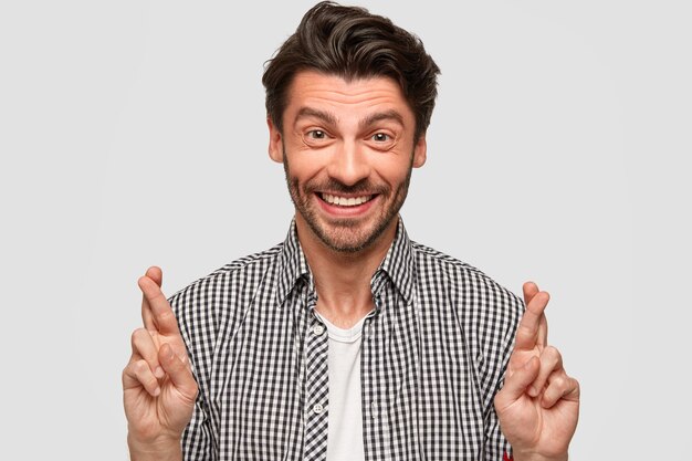 Happy Caucasian male with trendy haircut, gestures indoor, raises crossed fingers, believes in good luck, smiles sincerely, wears fashionable checkerd shirt, isolated over white wall. Expectation