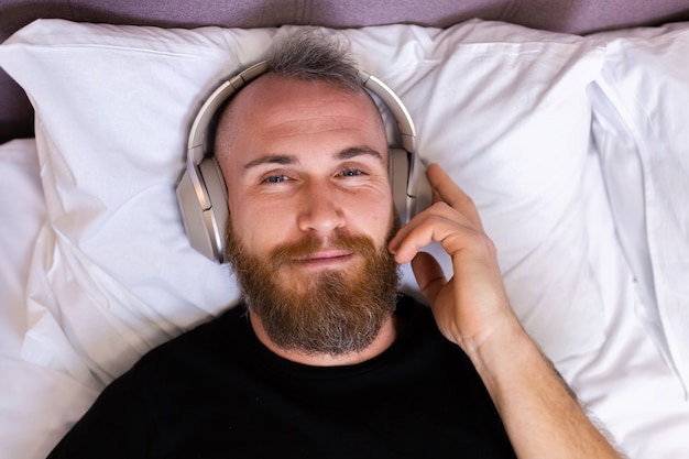 Happy caucasian man on bed wearing headphone listen enjoy his favorite music, resting alone, dancing.