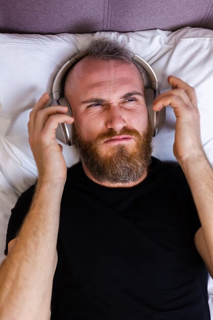 ヘッドフォンを身に着けているベッドの上の幸せな白人男性は、彼の好きな音楽を楽しんで、一人で休んで、踊っています。