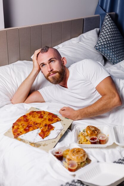 Happy caucasian man having fast food at home in bedroom on bed Man ordered food online take away and eat pizza and burgers in comfy room