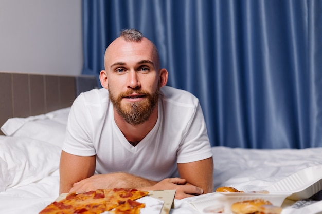 행복 한 백인 남자가 침대에 침실에서 집에서 패스트 푸드를 가지고 온라인으로 음식을 주문하고 편안한 방에서 피자와 햄버거를 먹습니다.