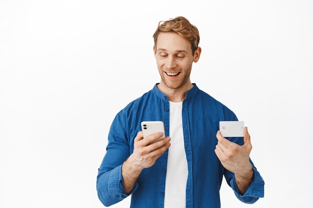 Happy caucasian man looking at smartphone while paying online with credit card, making order in mobile app, shopping in internet with bank card and cellphone, standing over white wall