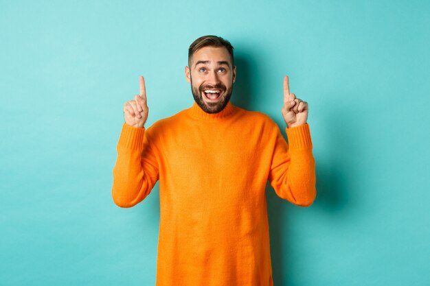 Happy caucasian man pointing fingers up, standing in sweater against turquoise wall.