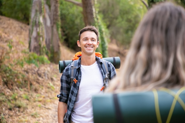 Happy Caucasian man standing in woods and smiling. Cheerful hiker walking in forest with blonde woman, enjoying nature, carrying backpack and posing. Tourism, adventure and summer vacation concept