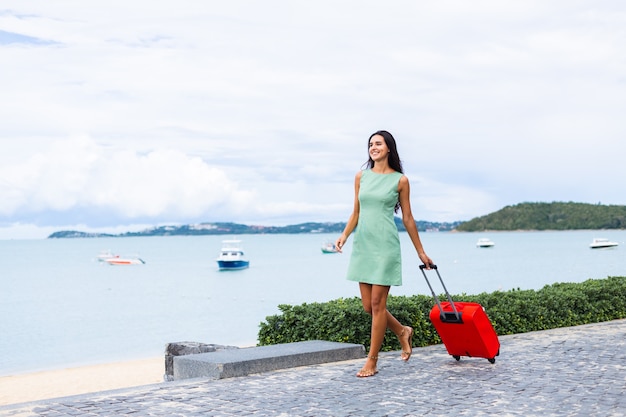Счастливая кавказская довольно длинными волосами, элегантная туристическая женщина в платье с красным чемоданом возле отеля