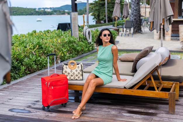 Happy caucasian pretty long hair elegant tourist woman in dress with red suitcase outside hotel