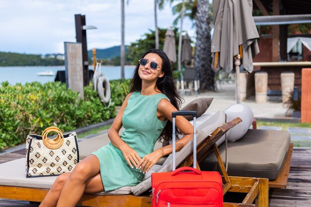 Happy caucasian pretty long hair elegant tourist woman in dress with red suitcase outside hotel