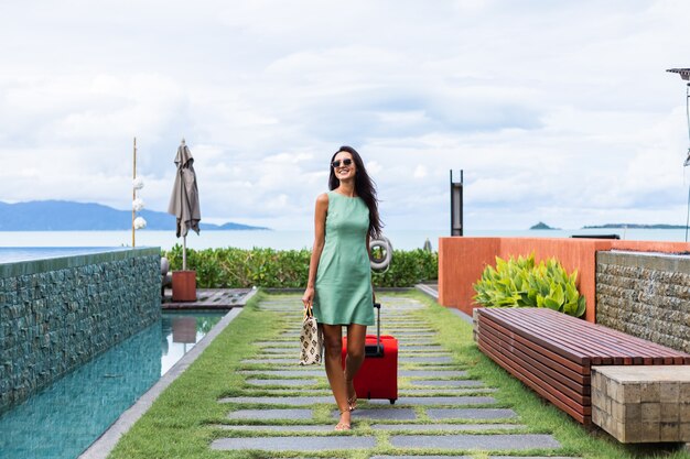 Happy caucasian pretty long hair elegant tourist woman in dress with red suitcase outside hotel