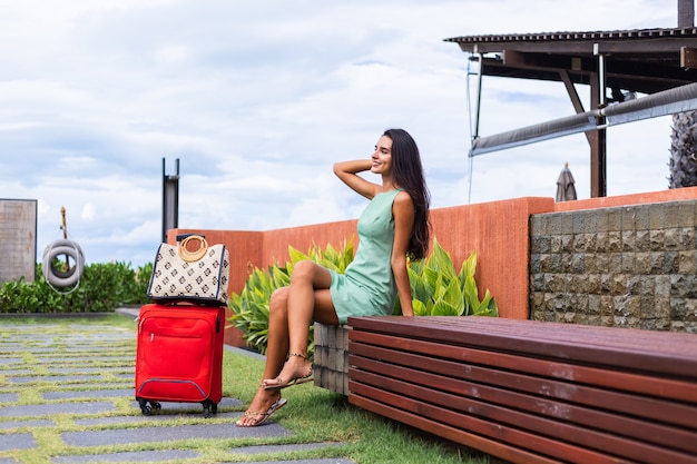 Счастливая кавказская довольно длинными волосами, элегантная туристическая женщина в платье с красным чемоданом возле отеля