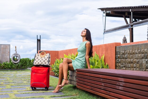 Happy caucasian pretty long hair elegant tourist woman in dress with red suitcase outside hotel