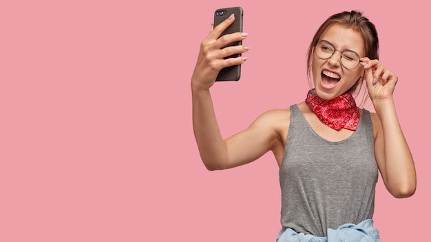 幸せな白人女性が目を瞬き、現代のスマートフォンで自分撮りのポーズをとる
