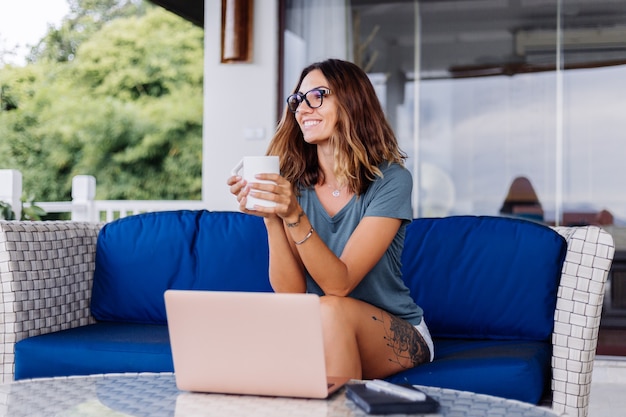 Happy caucasian woman does distance work on laptop at home comfortable place drinking tea