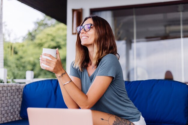 Happy caucasian woman does distance work on laptop at home comfortable place drinking tea