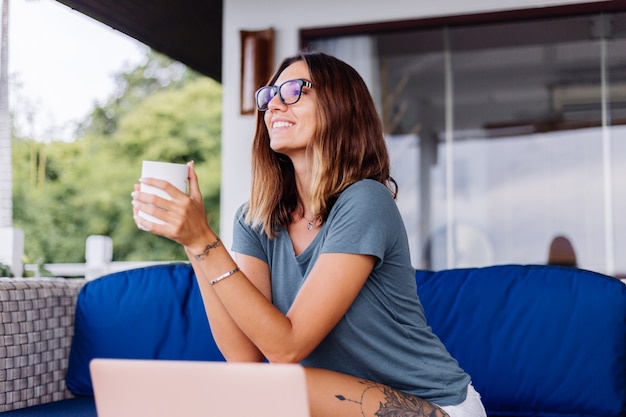 幸せな白人女性はお茶を飲む自宅の快適な場所でラップトップで遠隔作業を行います