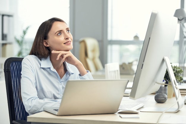 ノートパソコンと大画面の前のオフィスで夢を見て幸せな白人女性