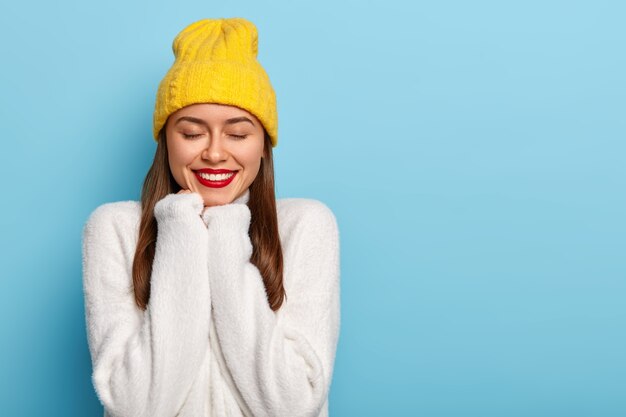 Happy Caucasian woman smiles pleasantly, has red painted lips, keeps hands under chin, wears cozy white winter sweater and yellow hat, keeps eyes closed, isolated over blue background, feels lucky