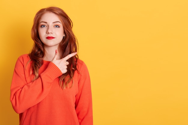 Happy Caucasian woman standing and pointing finger copy space on yellow space. Cute girl wearing casual orange sweater and finger pointing to aside for present promotions.