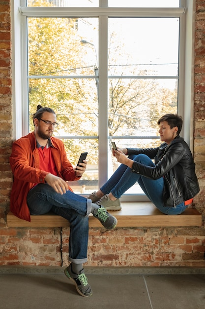 Happy caucasian young people, couple behind the brick window. sharing a news, photos or videos from smartphones, laptops or tablets, playing games and having fun. social media, modern technologies.