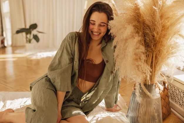 Free photo happy caucasian young woman laughing with her eyes closed while sitting on floor by window in living room brunette girl spends time alone on weekends modern house vacation concept