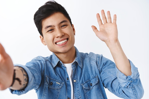 Happy charismatic asian man taking selfie on smartphone calling friends via videocall chat app waving hand to say hi hello communicating with followers via live stream white background