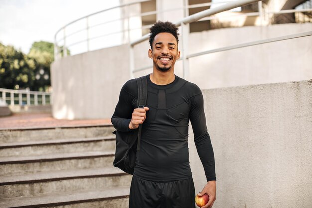 Happy charming brunette man in black sport outfit holds backpack and fresh apple Guy poses near stairs outside and smiles