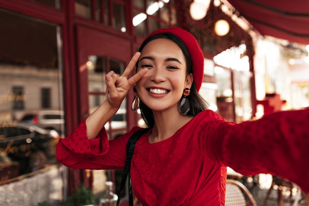 빨간 드레스, 세련된 베레모, 안경을 쓴 행복한 매력적인 브루네트 여성은 진심으로 미소를 지으며 평화의 표시를 보여주며 셀카를 밖에서 찍는다