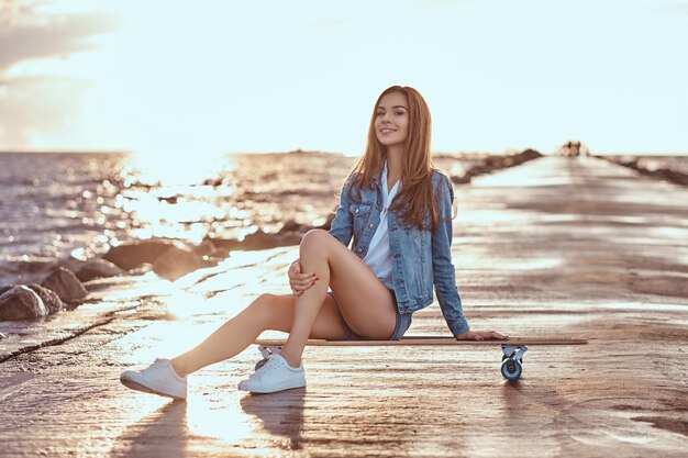 Счастливая очаровательная девушка, одетая в шорты и футболку, сидит на скейтборде на пляже во время яркого заката.