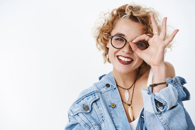 Happy charming girl staying positive looking through okay gesture feeling joyful and carefree being optimistic posing in glasses denim jacket over white background with stylish short curly hairstyle