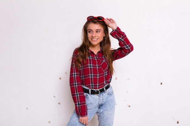 Happy charming girl wearing shirt and jeans in red glasses posing over isolated wall looking away and smiling