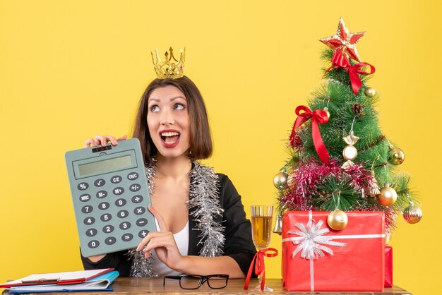 Happy charming lady in suit with crown holding calculator in the office on yellow isolated 