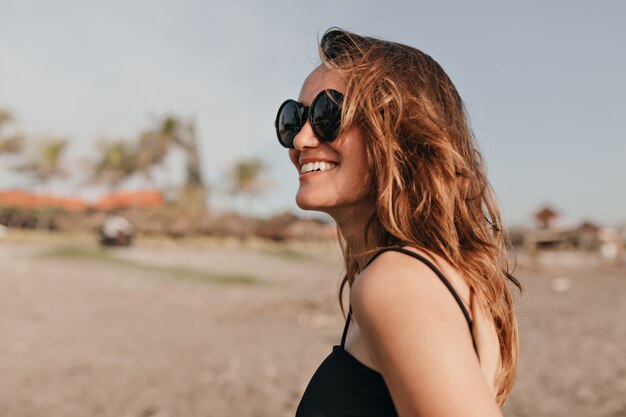 Happy charming smiling woman with dark wavy hair walking in sunshine on the beach and enjoying vacation on the ocean