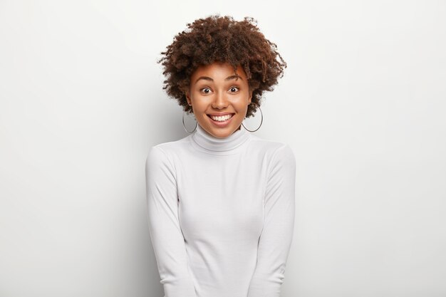 happy charming woman with afro hairstyle, wears snow white poloneck jumper, being in good mood after going shopping