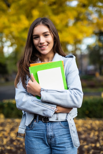 Happy charming young woman with backpack standing and holding notebooks in park