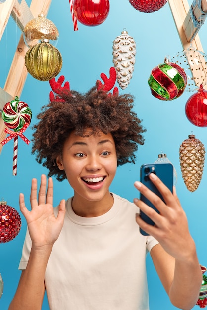 Happy cheerful African American woman waves hand at smartphone screen calls relatives stays at home during Christmas time enjoys cozy atmosphere decorates room before winter holidays. Festive mood