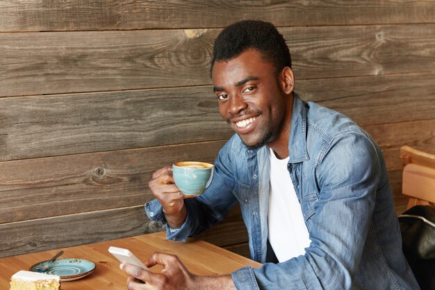 Happy cheerful African student holding mug, drinking fresh cappuccino, browsing internet and checking newsfeed on social media, using cell phone during coffee break at modern cafe with wooden walls