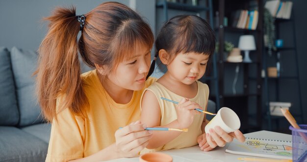 幸せな陽気なアジアの家族のお母さんは、家のリビングルームのテーブルでリラックスして楽しんでいる幼児の女の子のペイントセラミックポットを教えています