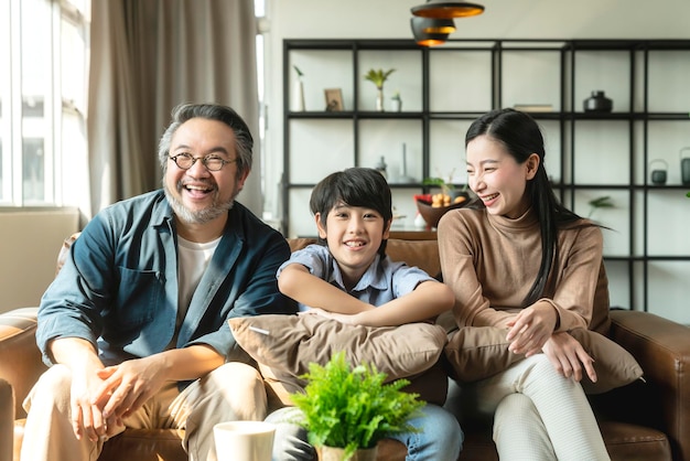 Happy cheerful Asian family parent with child boy son watching tv together spending holiday weekend time in quarantine on sofa in living room family and home concept